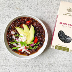 Women's Bean project black bean soup mix next to a bowl of the prepared soup topped with fresh avocado slices
