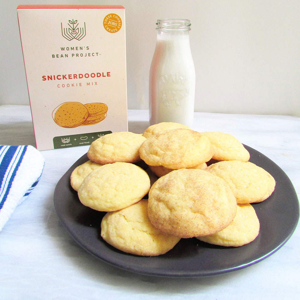 Women's Bean Project Snickerdoodle cookie mix behind a plate of fresh baked snickerdoodle cookies and a glass of milk