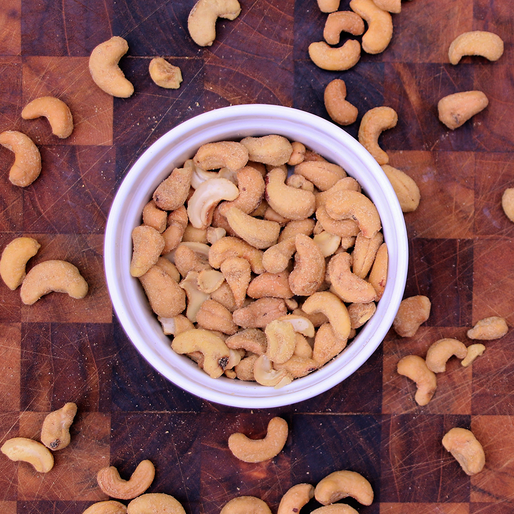 Women's Bean Project Thai Curry cashews in a bowl on a table surrounded by other cashews.