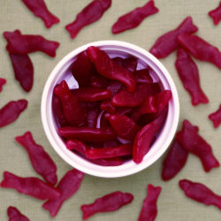 Women's Bean Project raspberry gummy fish in a white bowl on a table with gummy fish scattered around.