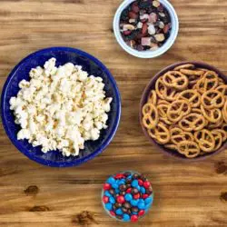 Four bowls on a table containing popcorn, chocolate candies, pretzels, and trail mix.