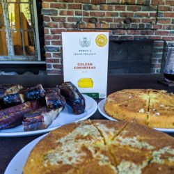 Women's Bean Project cornbread mix box next to a plate of barbecue ribs and baked cornbread.